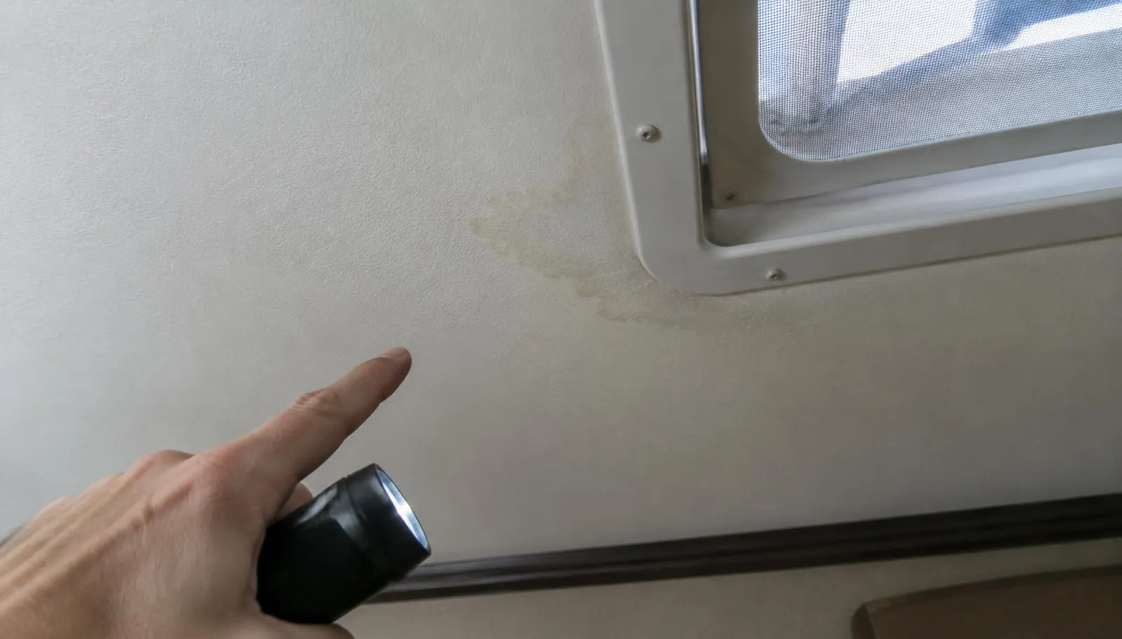 Hand with a flashlight pointing to a faint water stain on an RV ceiling near a roof vent